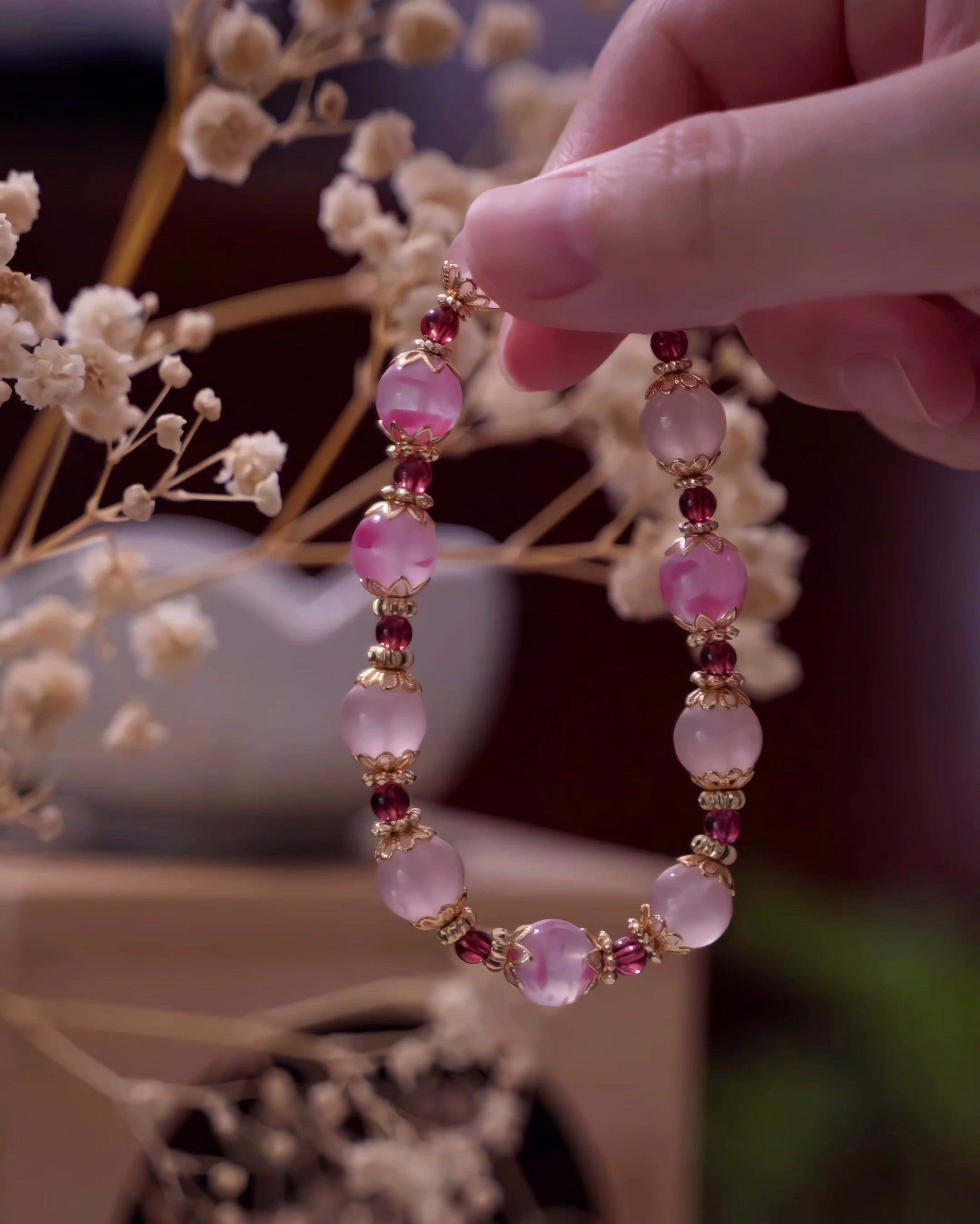 Hand holding a bracelet with pink beads and gold accents against a blurred natural background